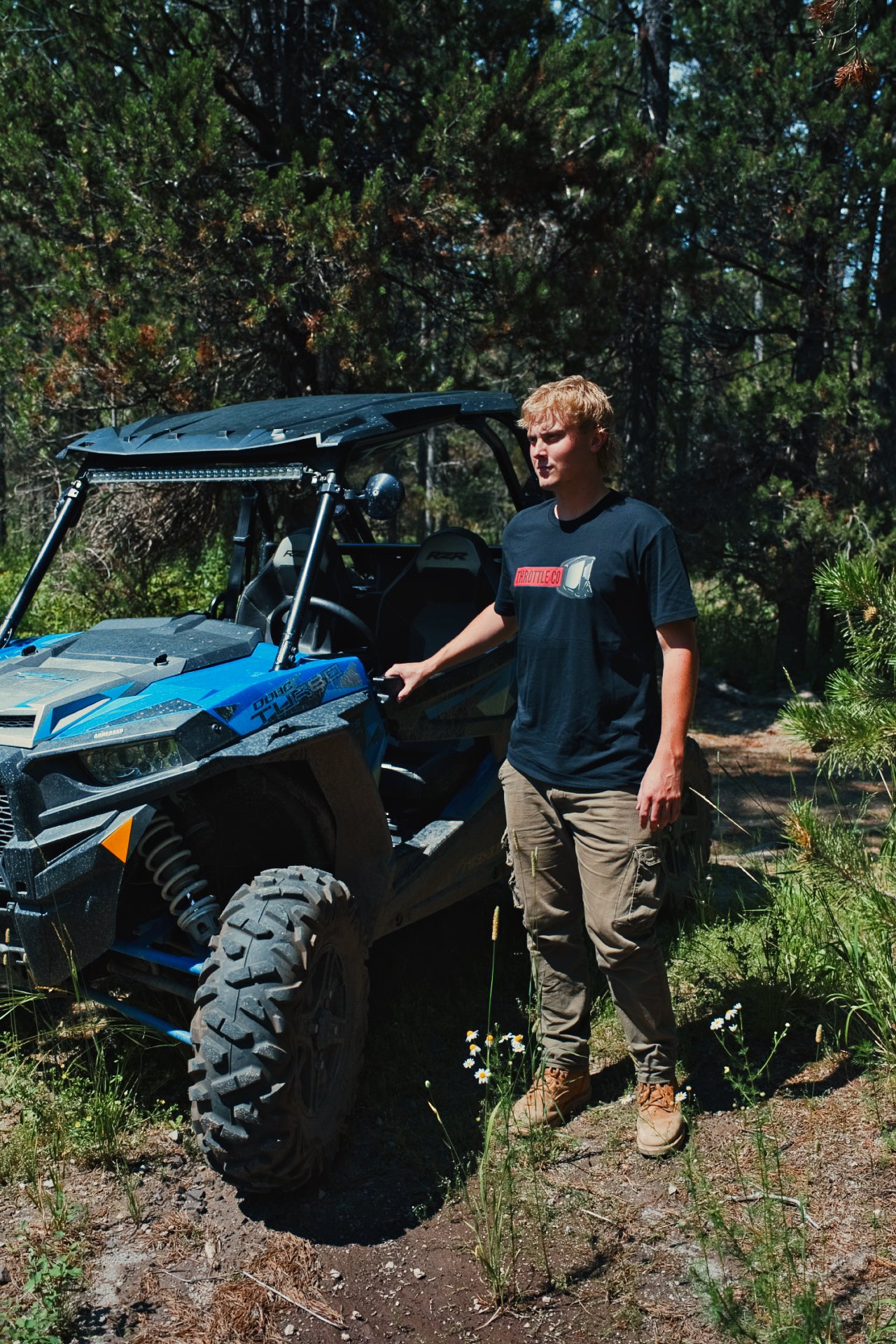 Rider in the Throttle Co Red Goggles Tee standing by a blue UTV on a forest trail