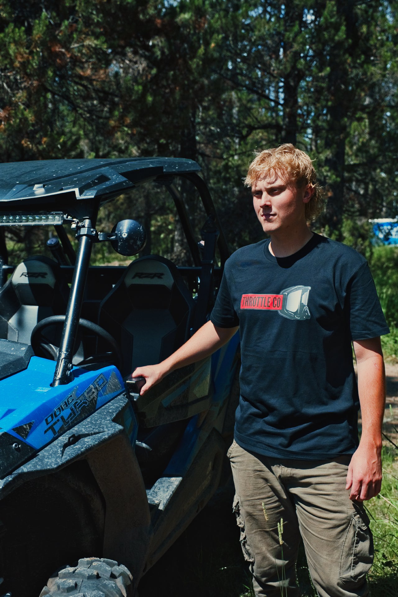 Rider wearing the Throttle Co Red Goggles Tee standing beside a blue UTV in the trees