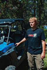 Rider wearing the Throttle Co Red Goggles Tee standing beside a blue UTV in the trees
