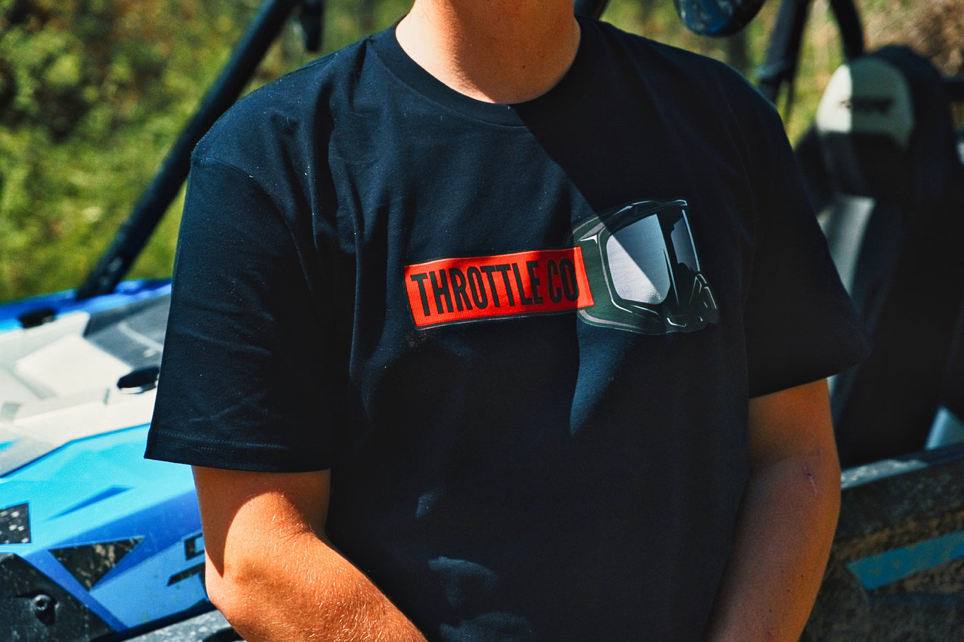 Close-up of the red Throttle Co badge and goggles chest graphic, blue UTV in the background