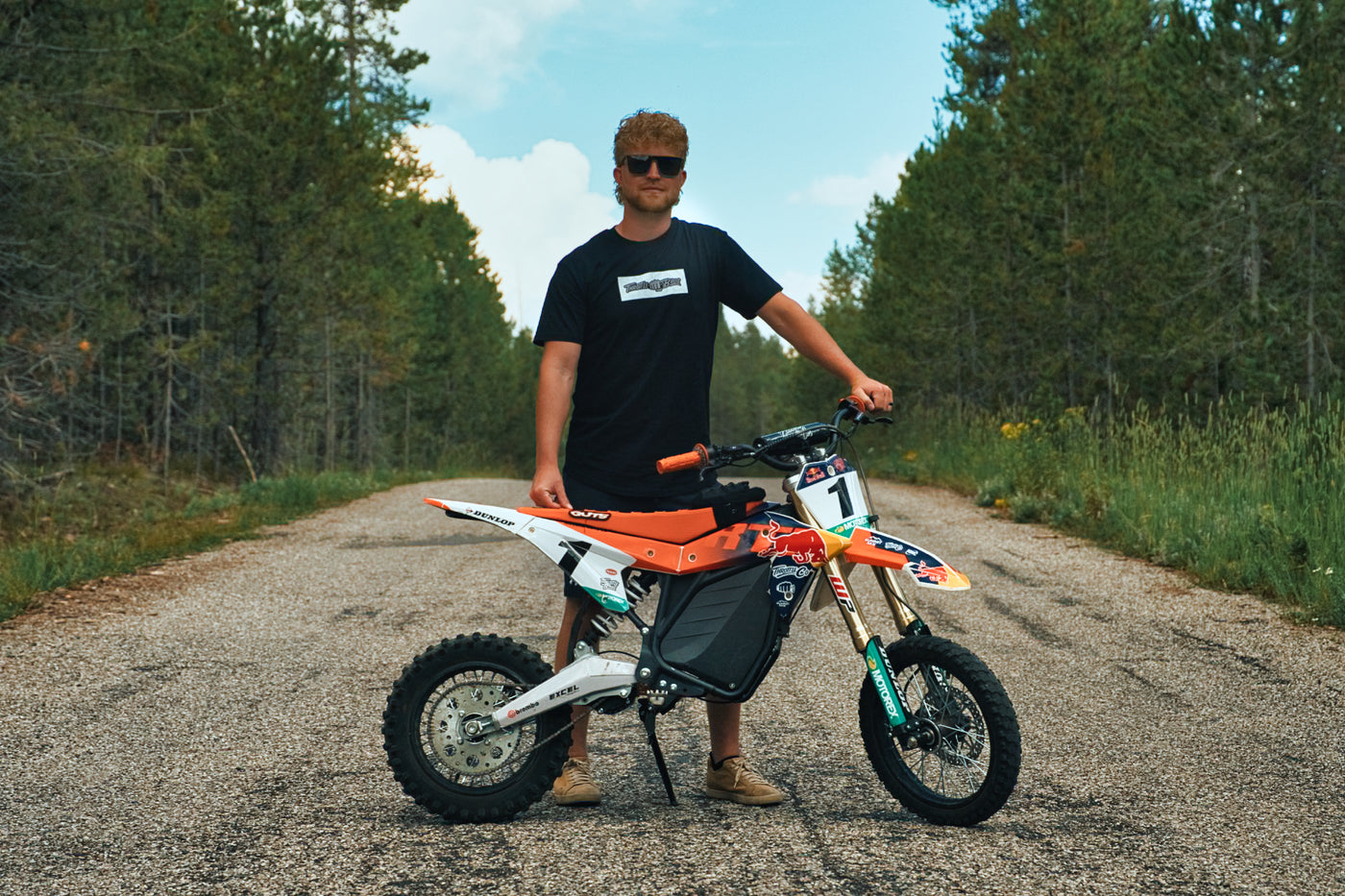 Rider in the Throttle Bros Logo Topo Tee standing behind a dirt bike on a tree-lined gravel road