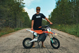 Rider in the Throttle Bros Logo Topo Tee standing behind a dirt bike on a tree-lined gravel road
