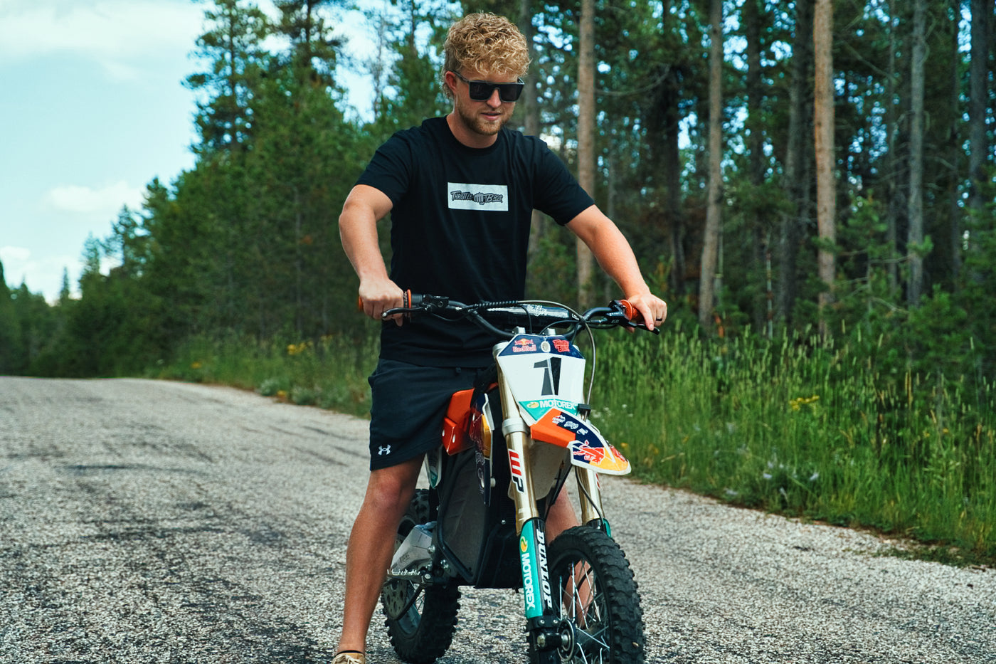 Rider wearing the Throttle Bros Logo Topo Tee standing over a dirt bike on a forest road