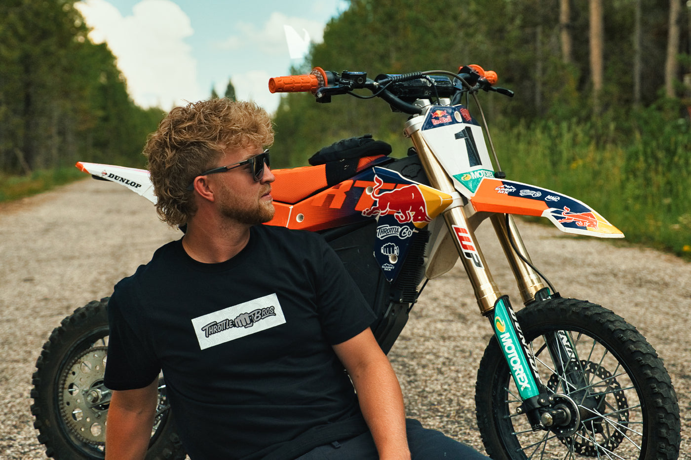 Rider in the Throttle Bros Logo Topo Tee crouched beside a dirt bike on a forest gravel road