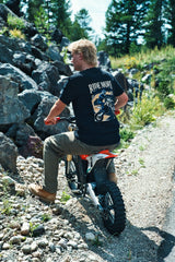 Rider seated on a dirt bike on a rocky trail wearing the Ride More Bike Tee, back graphic visible