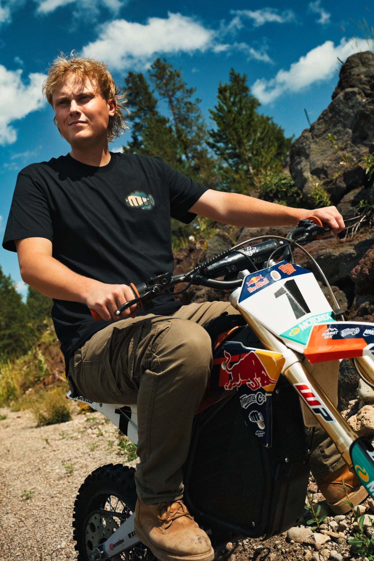 Rider standing with a dirt bike among rocks and trees in the black Ride More Bike Tee