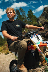 Rider standing with a dirt bike among rocks and trees in the black Ride More Bike Tee