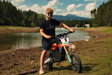 Rider in the black Topo Design Tee standing over a dirt bike with a lake and mountains behind
