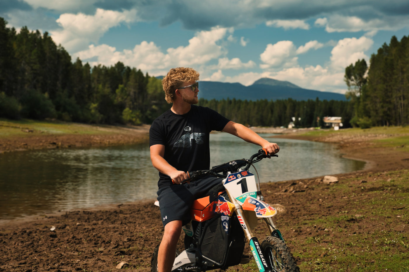 Rider wearing the black Topo Design Tee standing behind a dirt bike at a mountain lake