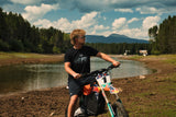 Rider wearing the black Topo Design Tee standing behind a dirt bike at a mountain lake