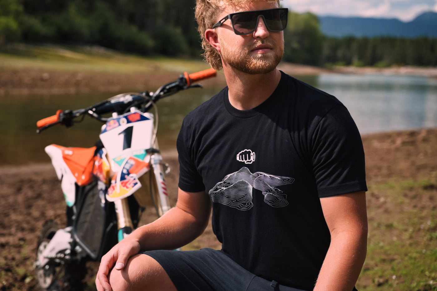 Rider in the black Throttle Co Topo Design Tee crouched beside an orange and white dirt bike on a lakeshore