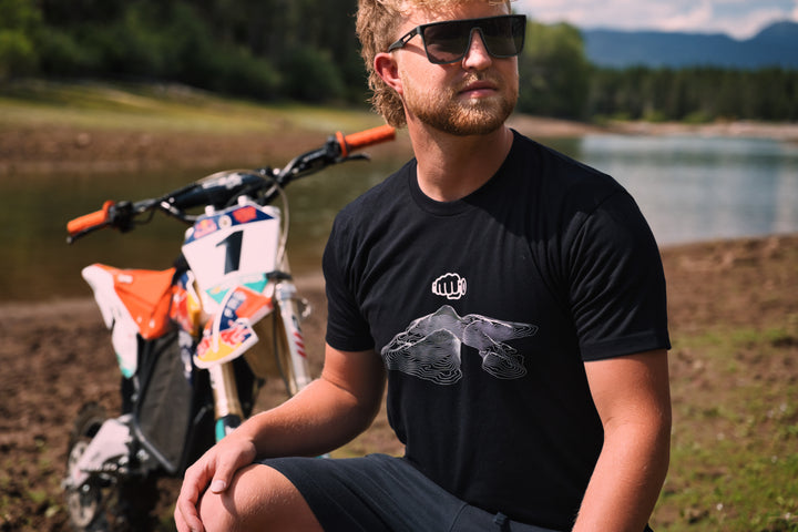 Rider in the black Throttle Co Topo Design Tee crouched beside an orange and white dirt bike on a lakeshore