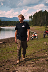 Rider in the Fist Icon Topo Design tee walking the shoreline with a dirt bike and UTV parked behind