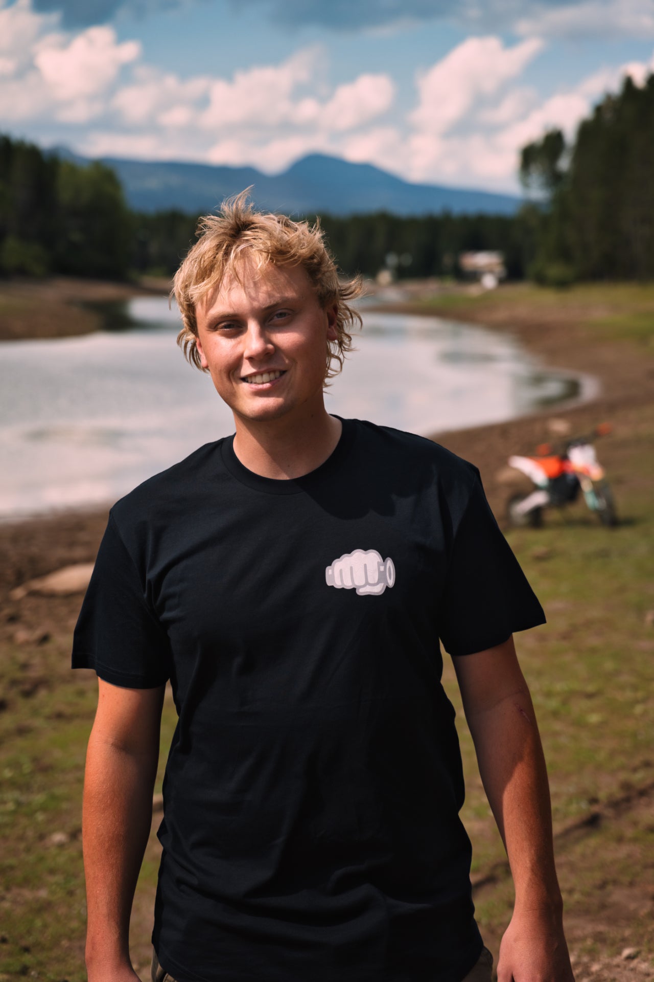 Rider wearing the black Fist Icon Topo Design tee at a mountain lakeshore