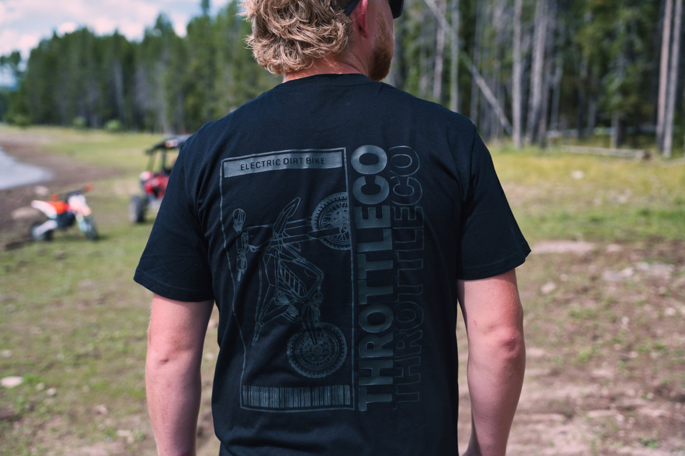 Rider wearing the Throttle Co Electric Dirt Bike Barcode Tag Tee in black, back view showing the tonal ELECTRIC DIRT BIKE tag graphic with line-art dirt bike, barcode and stacked THROTTLECO logo, shot trailside with a dirt bike and UTV