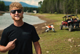 Rider pointing at the tonal blackout THROTTLECO chest logo on the black Electric Dirt Bike Barcode Tag Tee, lakeside with a dirt bike and UTV behind him