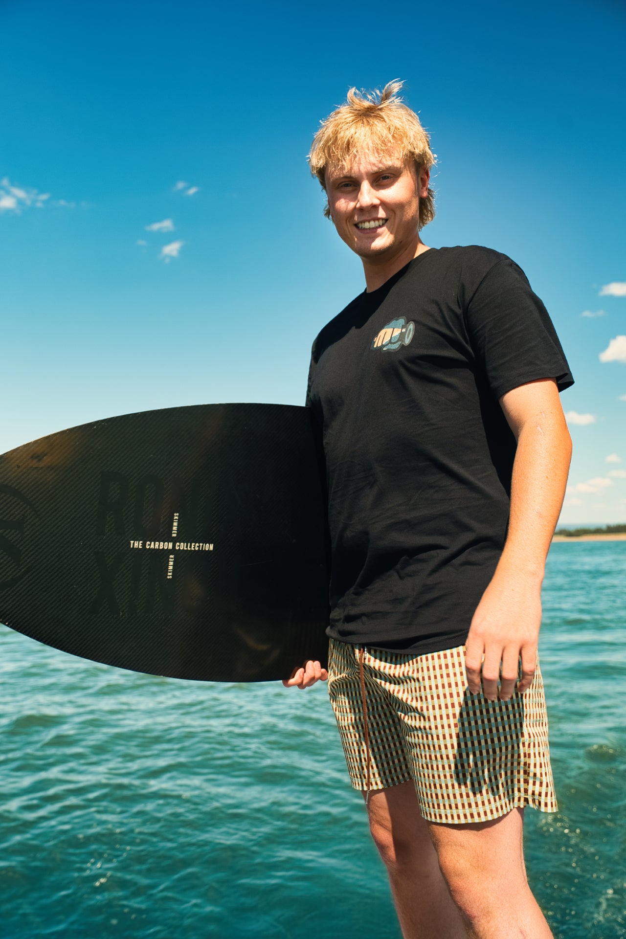 Rider holding a wakeboard by the lake in the black Ride More Bike Tee, front chest graphic visible