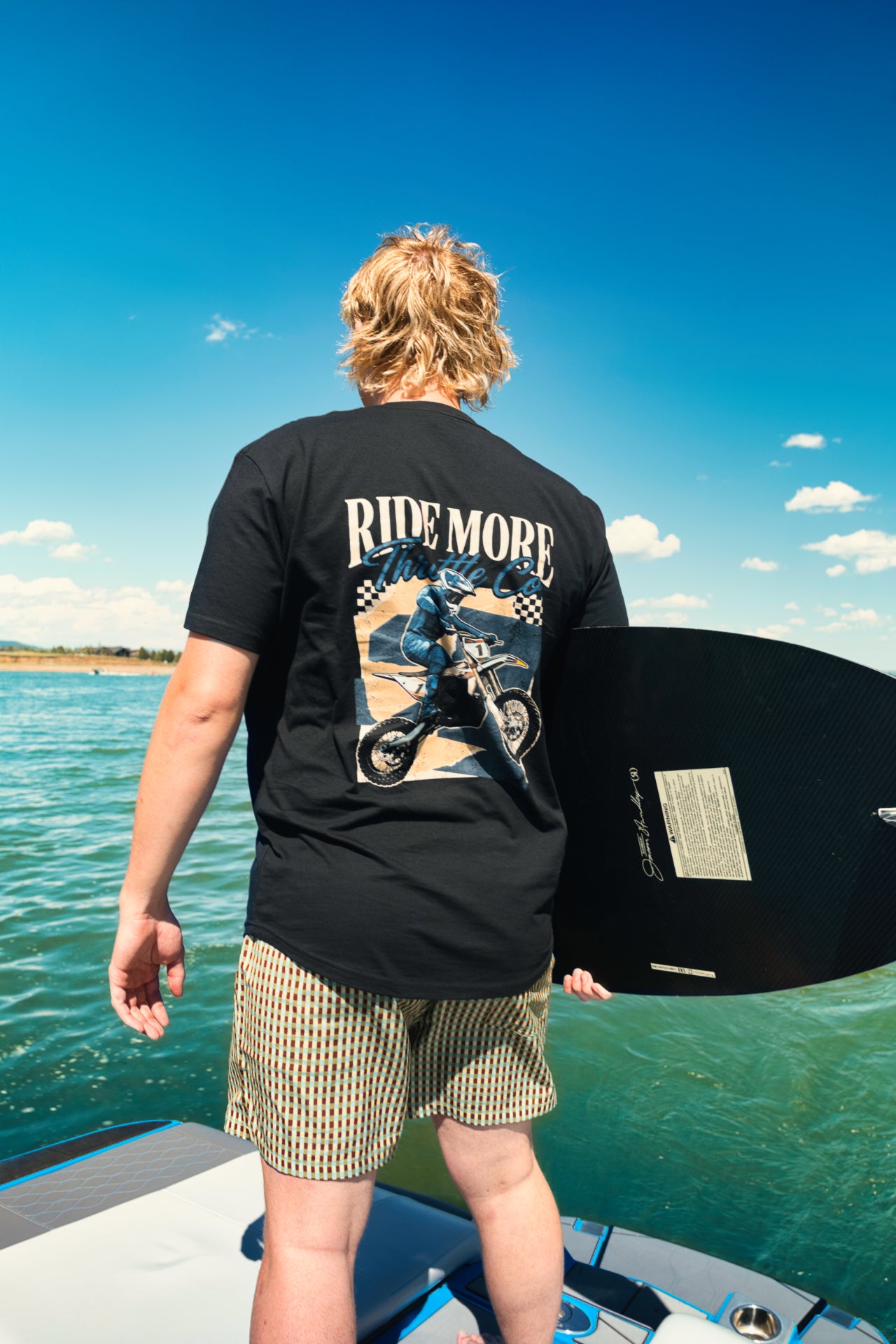 Rider on a boat holding a wakeboard, wearing the Ride More Bike Tee with the RIDE MORE back graphic visible