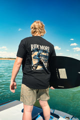 Rider on a boat holding a wakeboard, wearing the Ride More Bike Tee with the RIDE MORE back graphic visible