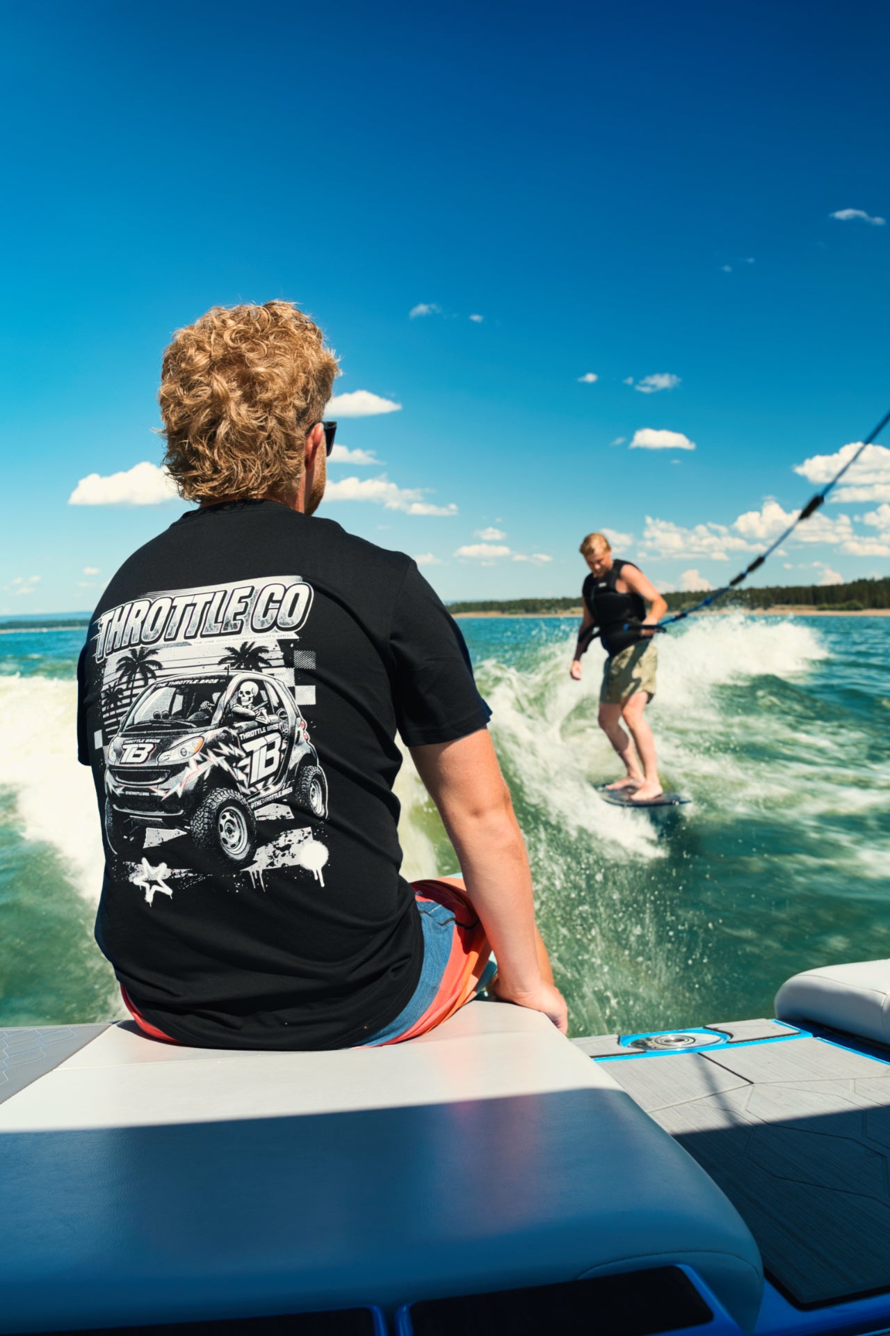 Rider on a wake boat in the Smart Car Throttle Bros Tee watching a wakesurfer, back graphic visible
