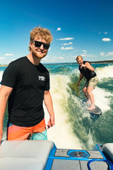 Rider standing on a wake boat in the Smart Car Throttle Bros Tee with a wakesurfer riding the wake behind